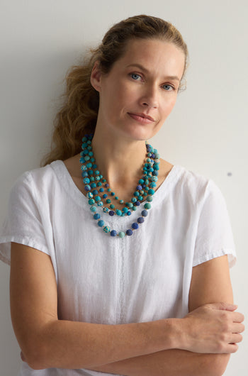 A woman in a white shirt stands against a simple light background, wearing the Kantha Cascade Necklace made from repurposed fabric beads.