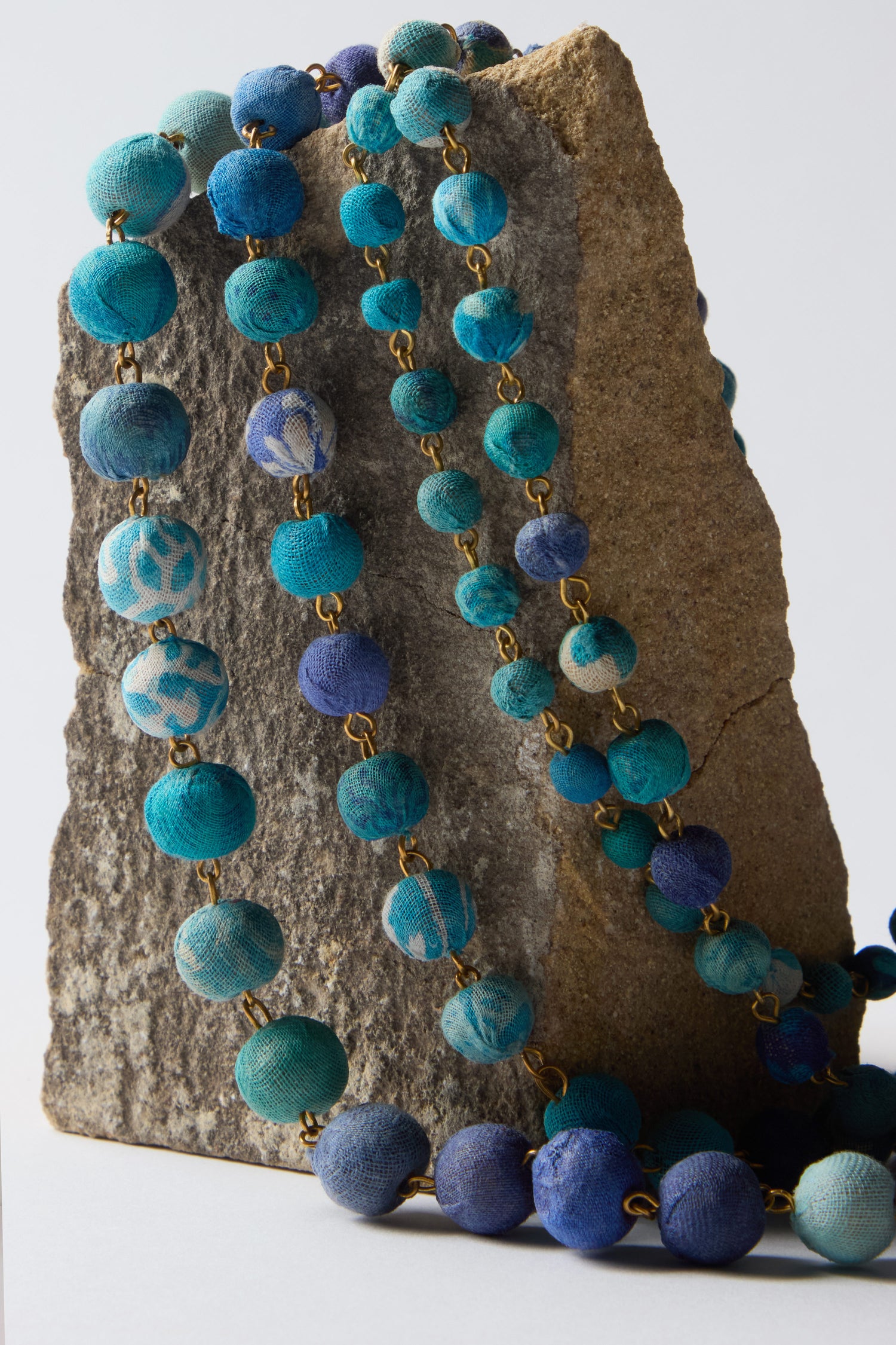 The Kantha Cascade Necklace, featuring vibrant blue and turquoise repurposed fabric beads, elegantly drapes over a textured rock, showcasing its dedication to sustainable fashion.