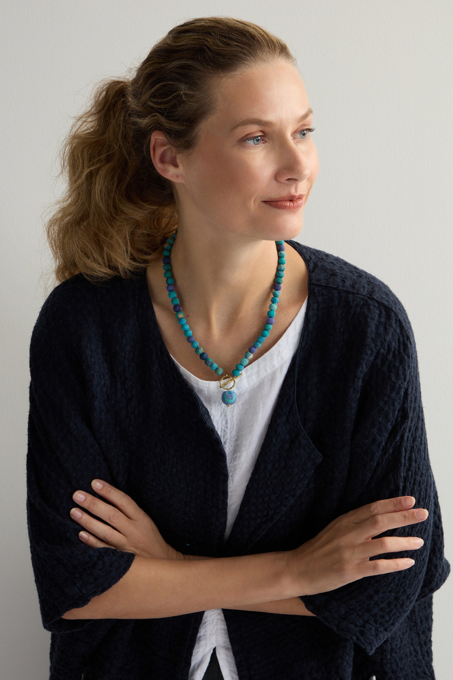 A woman with curly hair, wearing a handcrafted Kantha Toggle Necklace, black cardigan, and white top, stands with arms crossed, looking to the side.