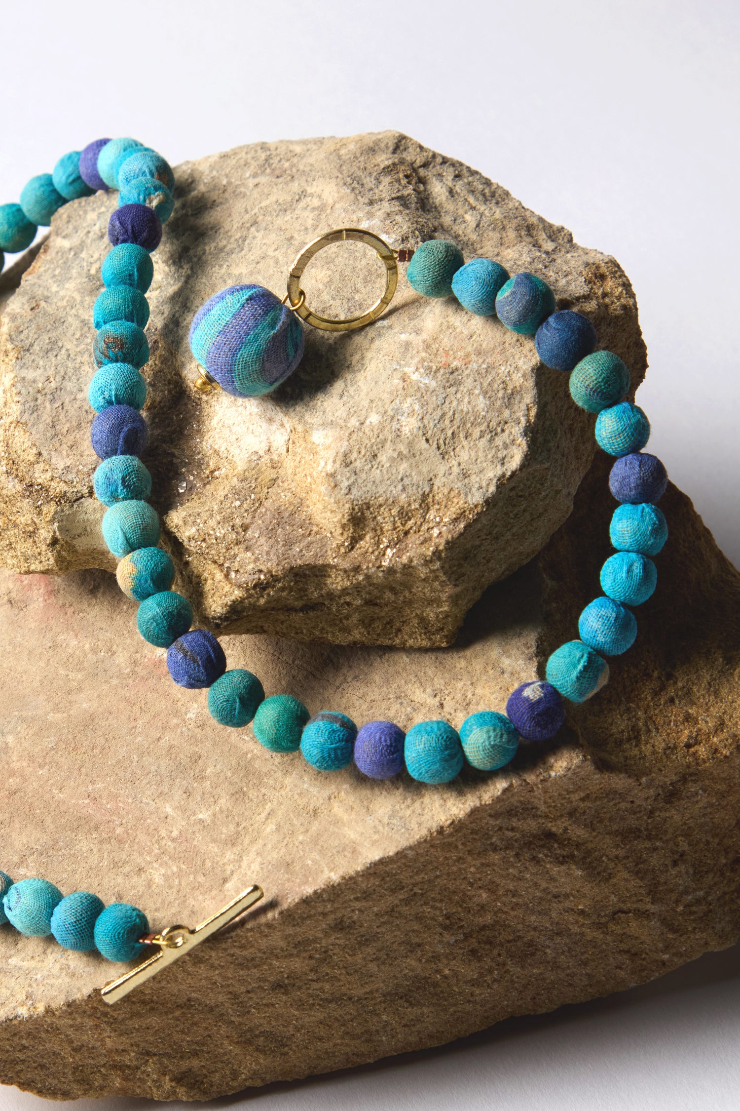 The Kantha Toggle Necklace, showcasing artisanal craftsmanship, features turquoise and navy beads with a gold clasp and is elegantly displayed on a textured rock surface.