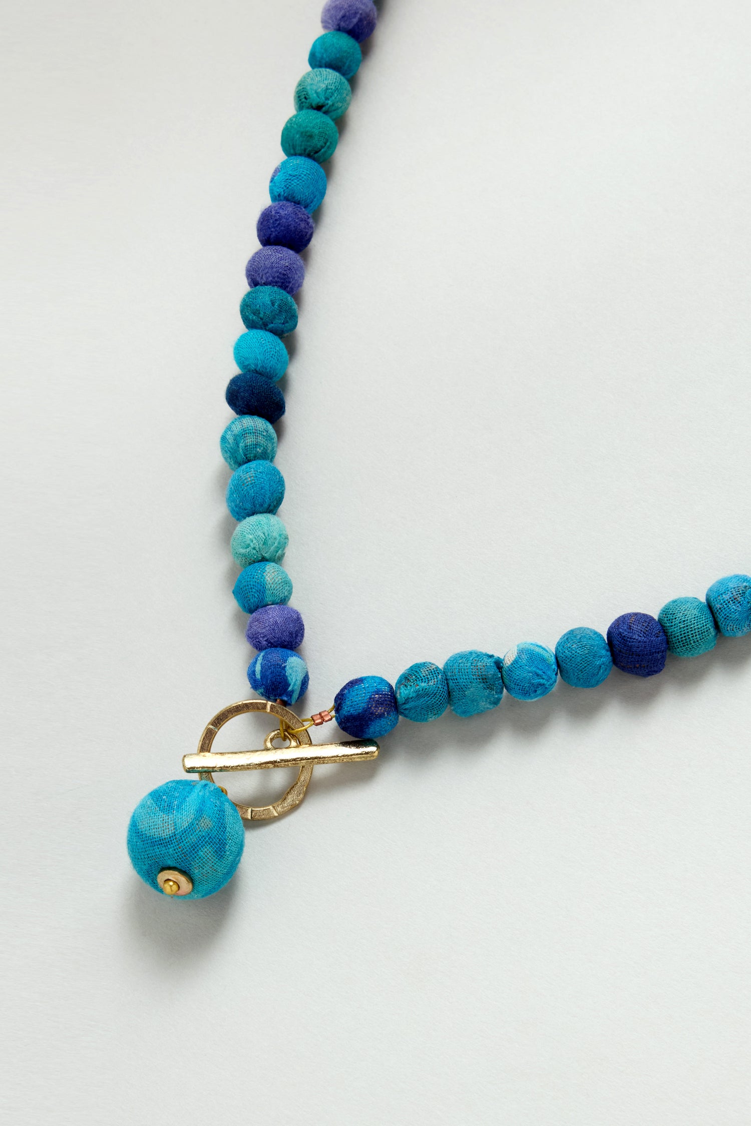 The Kantha Toggle Necklace features small blue fabric-covered beads made from repurposed textiles, a bold larger blue pendant, and a gold toggle clasp, all showcased against a light background.