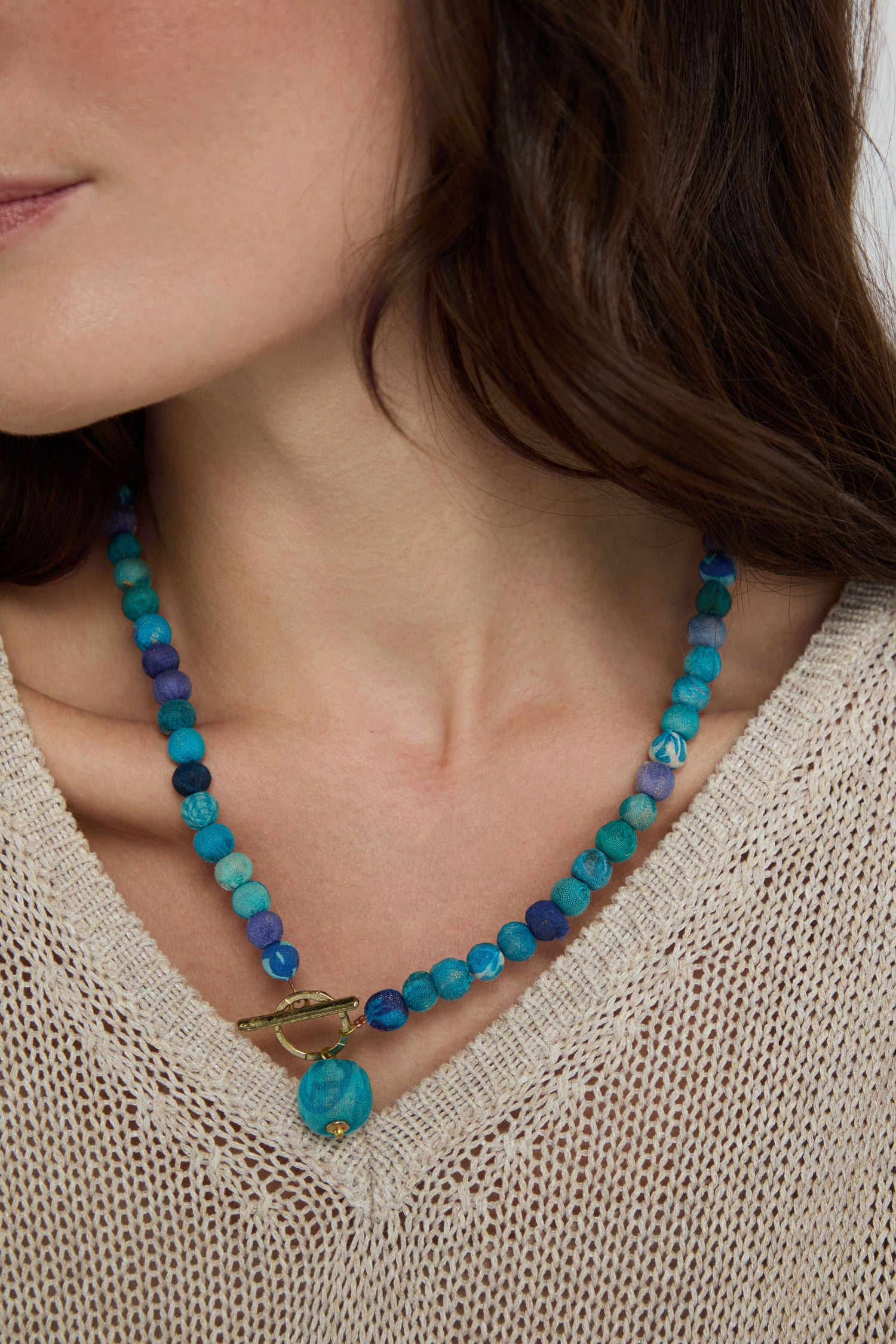 A woman wears a beige knit top and a blue Kantha Toggle Necklace made from repurposed textiles, featuring a gold toggle clasp.