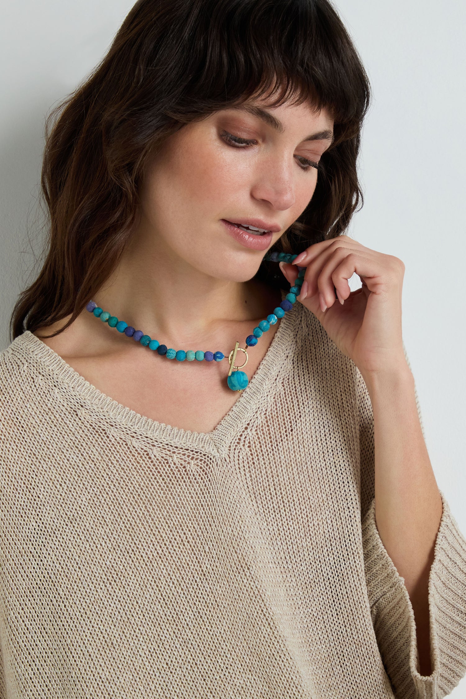 A woman in a beige knit top looks down, holding the Kantha Toggle Necklace—artfully handmade from upcycled textiles—in one hand.