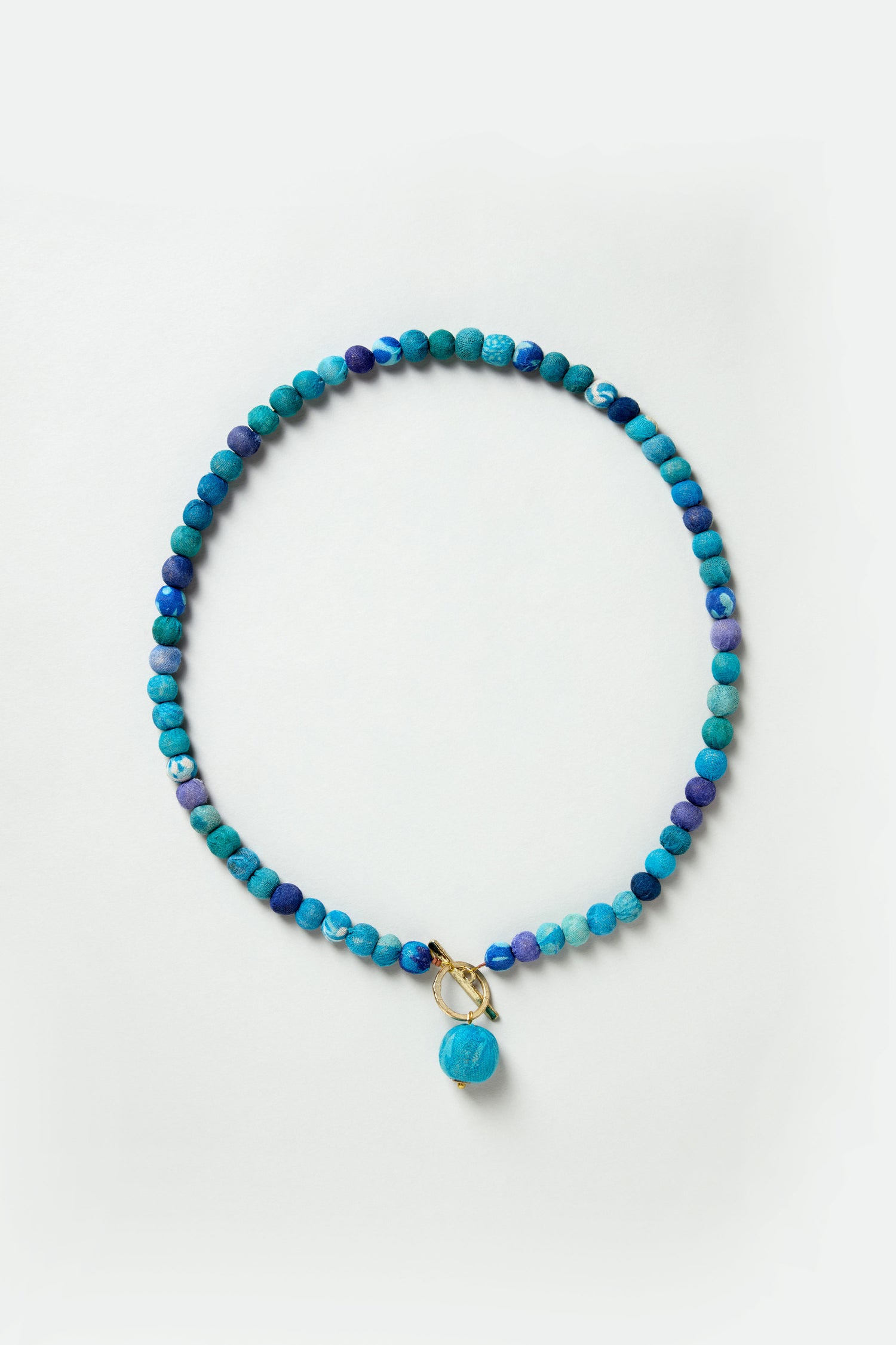 The Kantha Toggle Necklace features round blue stones, a gold-toned clasp, and a central blue charm, beautifully displayed on a light background.