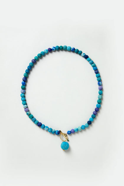 The Kantha Toggle Necklace features round blue stones, a gold-toned clasp, and a central blue charm, beautifully displayed on a light background.