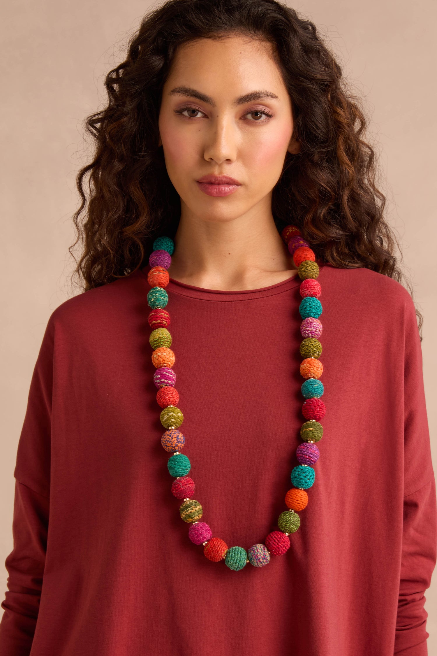 A woman with curly hair wears the Rainbow Kantha Spheres Necklace—featuring multicolored, cotton-wrapped beads—paired with a loose red top, standing against a beige background.