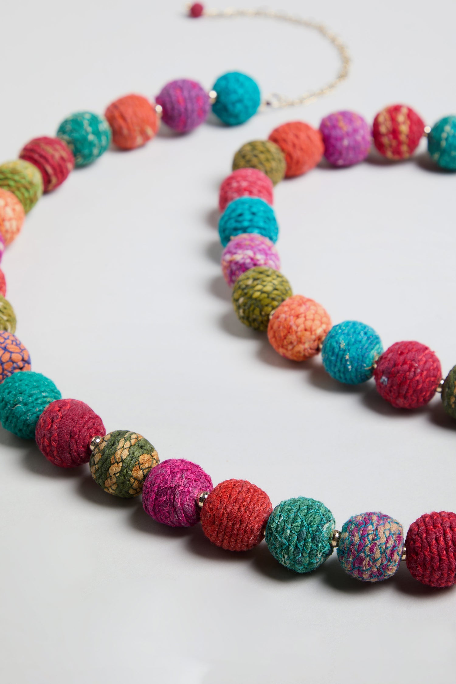 The Rainbow Kantha Spheres Necklace showcases round, textured cotton-wrapped beads in vibrant shades of red, green, blue, orange, and purple, displayed on a light background—a striking piece of colorful jewelry.