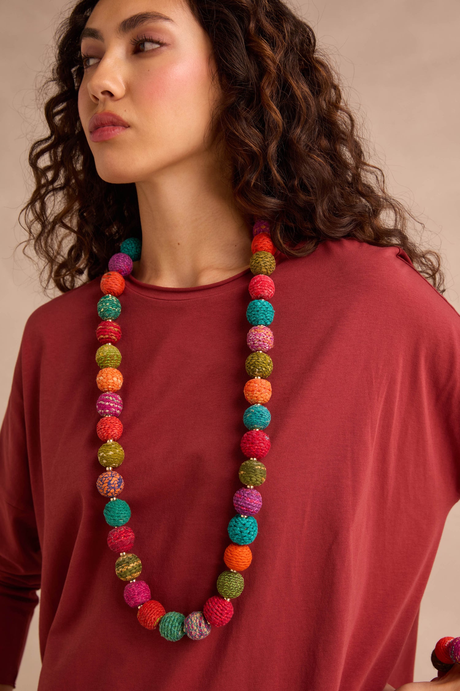 A woman stands against a neutral background wearing a loose-fitting maroon top and the Rainbow Kantha Spheres Necklace, featuring long strands of colorful, cotton-wrapped beads.