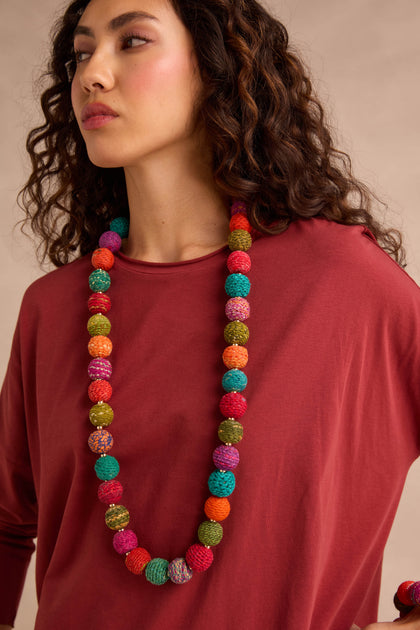 A woman stands against a neutral background wearing a loose-fitting maroon top and the Rainbow Kantha Spheres Necklace, featuring long strands of colorful, cotton-wrapped beads.
