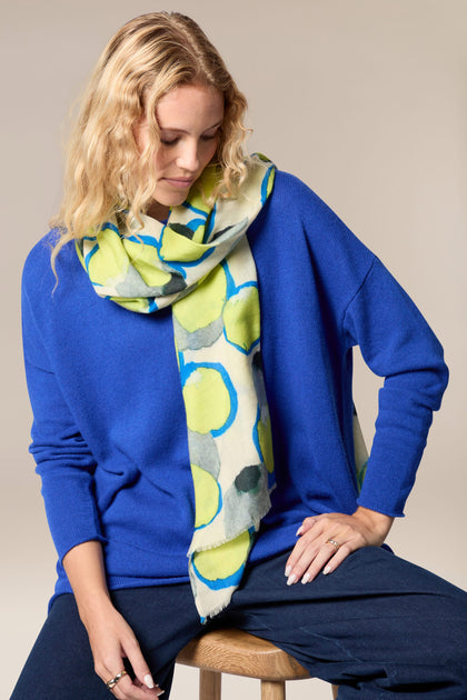 A woman is sitting on a stool wearing a blue sweater and a Neon Spot Wool Scarf with all-occasion styling.