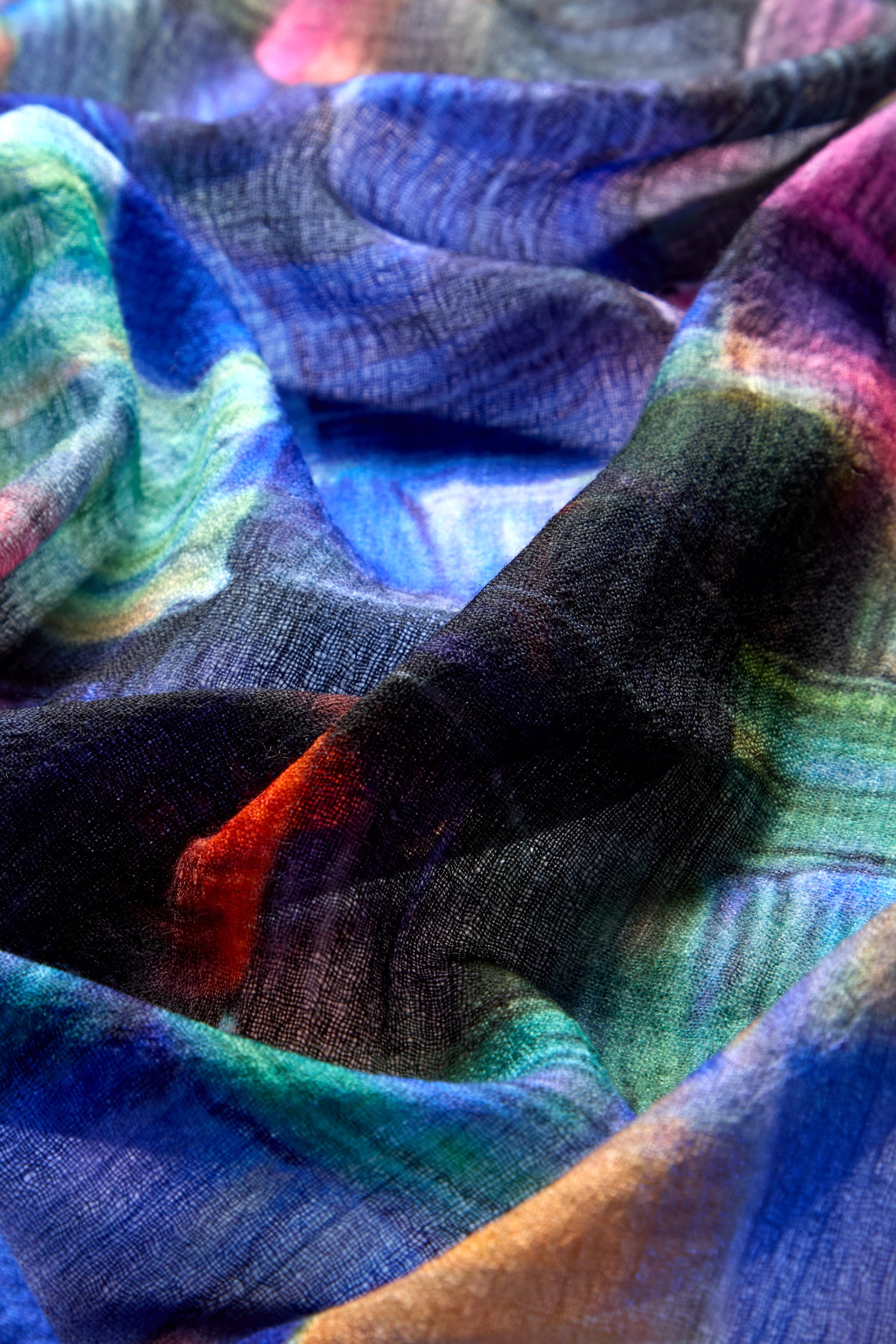 A close-up of the Painterly Merino Wool Scarf showcases a multi-colored, wrinkled fabric with hues of blue, green, purple, and black, reminiscent of wearable art.