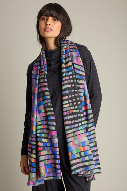 A person wearing a dark outfit and a Graduated Rainbow Squares Wool Scarf stands against a plain background.