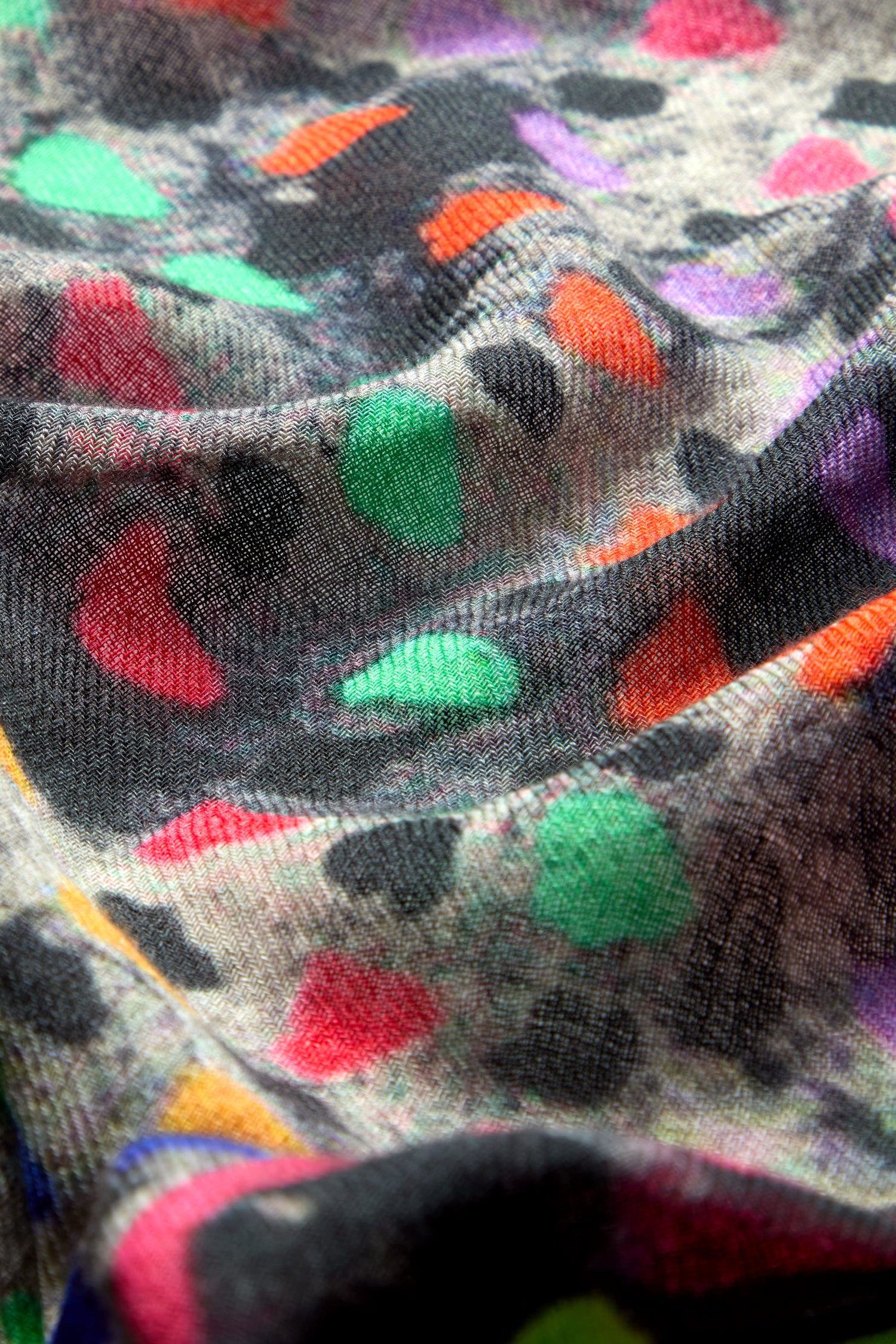 Close-up of the Painted Triangles Wool Scarf, showcasing its high-quality wool fabric adorned with a multicolored, abstract pattern. The scarf features vibrant red, green, purple, and orange shapes on a gray and black background, resembling a rainbow triangle print. The design reveals a textured and slightly wrinkled surface.