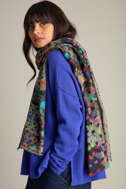 A person stands with the vibrant **Painted Triangles Wool Scarf** draped over their shoulder, wearing a bright blue sweater and dark pants, looking toward the camera. The high-quality wool scarf features an artistic rainbow triangle print that adds a touch of elegance to their outfit.