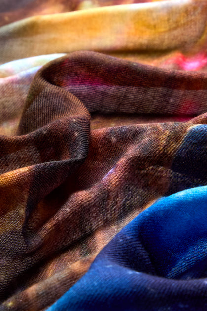 Close-up of a colorful, flowing fabric with a mix of brown, blue, and yellow hues. The texture appears soft and slightly transparent, embodying celestial elegance. This **Cosmic Sky Print Wool Scarf** exudes a captivating charm in every detail.