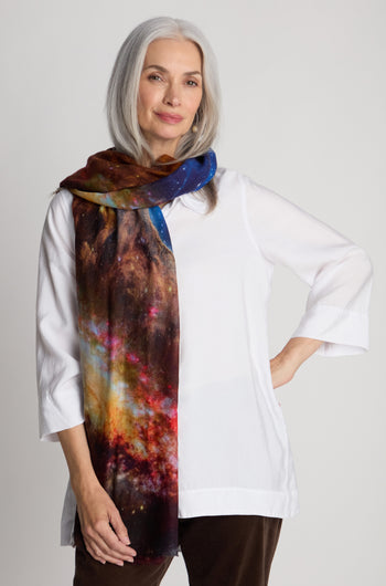 A woman with long gray hair wears a white blouse and a wool scarf featuring a cosmic sky print, standing against a plain background.