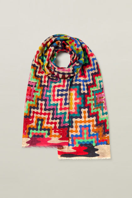 A vibrant scarf featuring a psychedelic wavy design with zigzags and dots in an array of red, blue, green, yellow, and pink hues. Crafted from premium soft wool, the ends of this Reverie Print Wool Scarf are artistically frayed. It lies flat on a light grey surface.