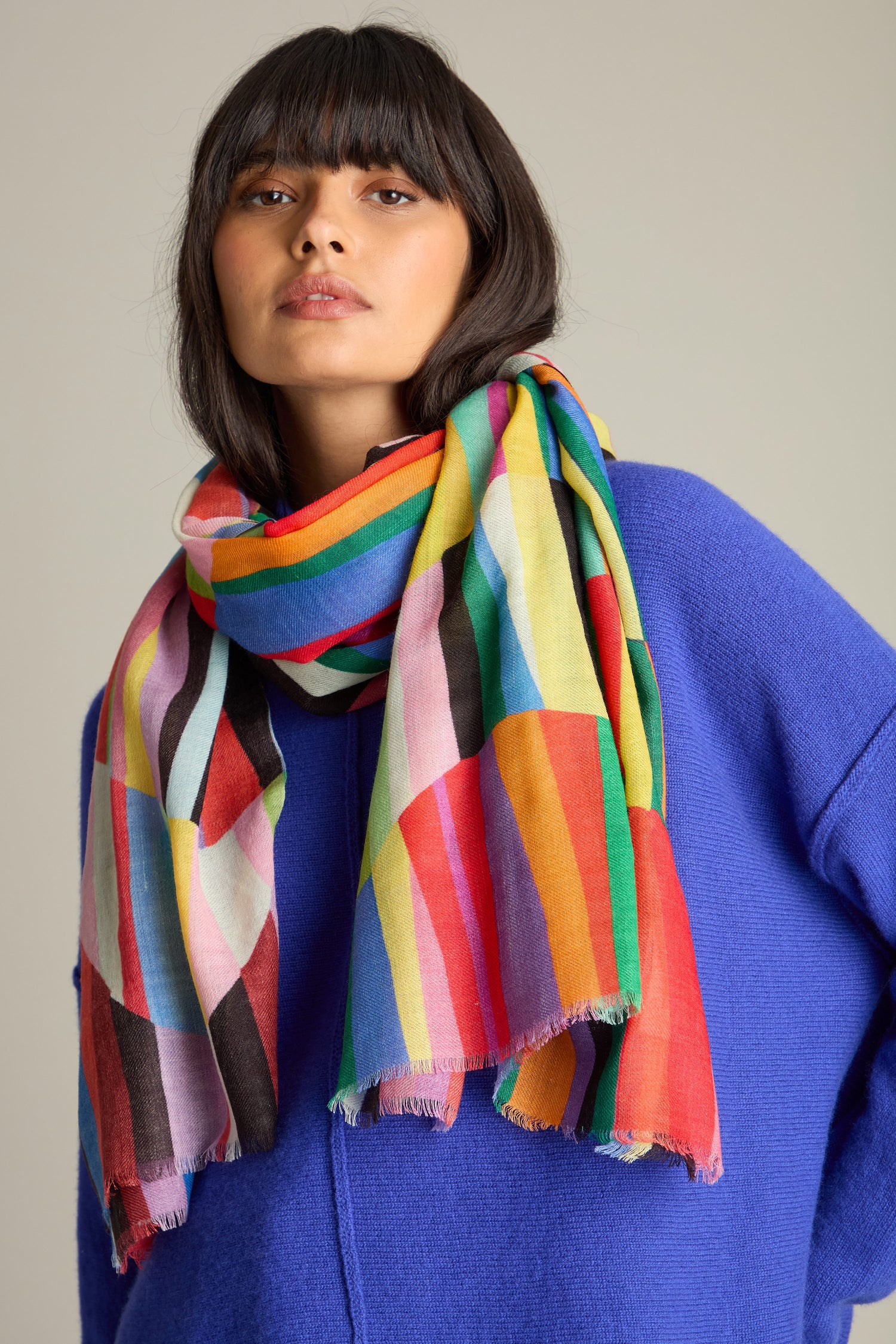 A person wearing a blue sweater and the Geometric Rainbow Stripe Wool Scarf looks directly at the camera.