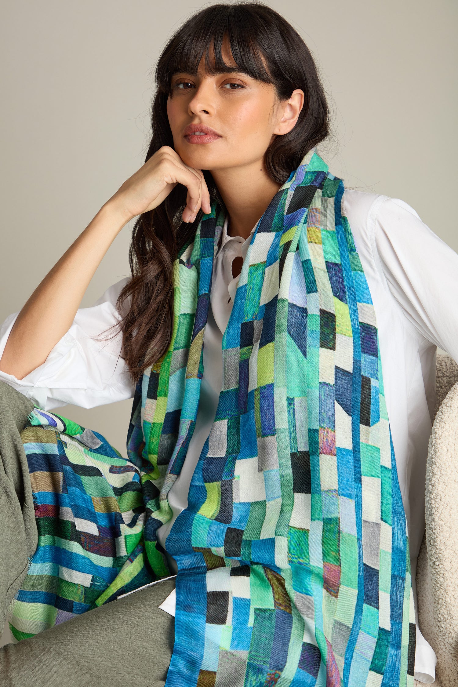 A woman with long dark hair poses while wearing a chic Ocean Squares Wool Scarf over a white shirt and green pants.