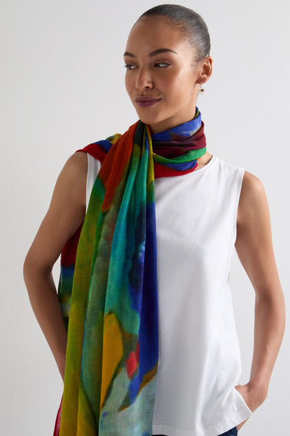 Against a plain background, someone showcases versatile styling by wearing a white sleeveless top paired with the vibrant Painterly Wool Blend Scarf.