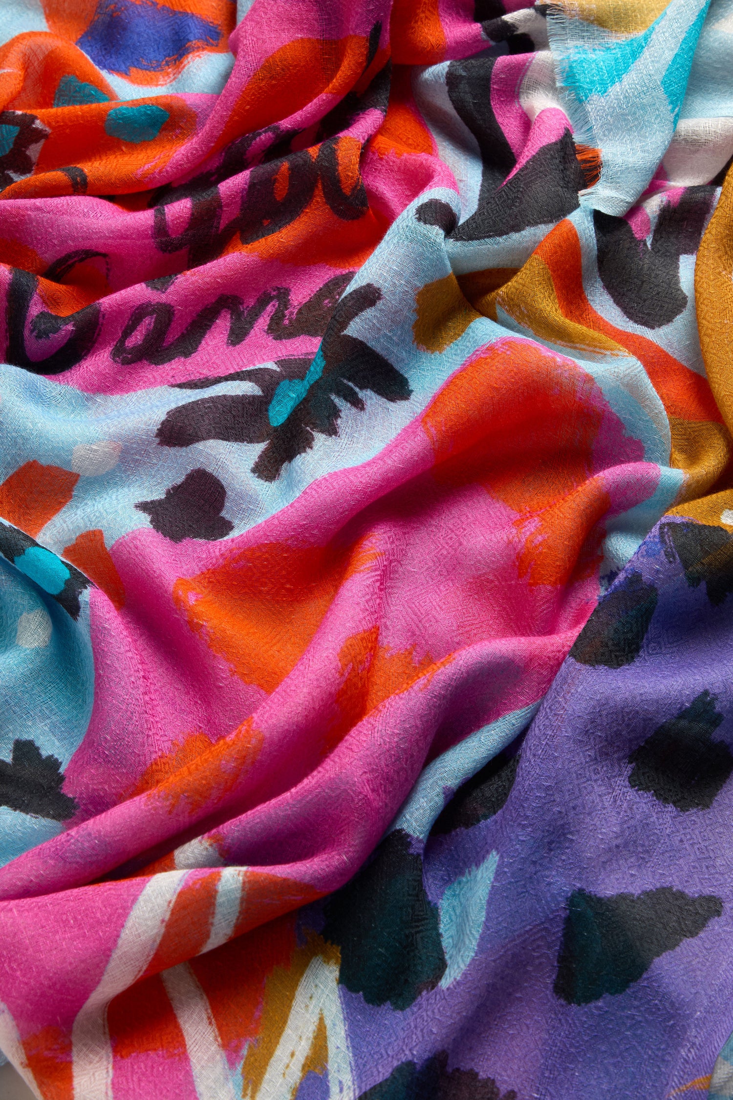 The Painted Stars Cotton Blend Scarf displays abstract patterns in pink, blue, orange, and purple with partially visible black text and an artistic star motif for a celestial touch.