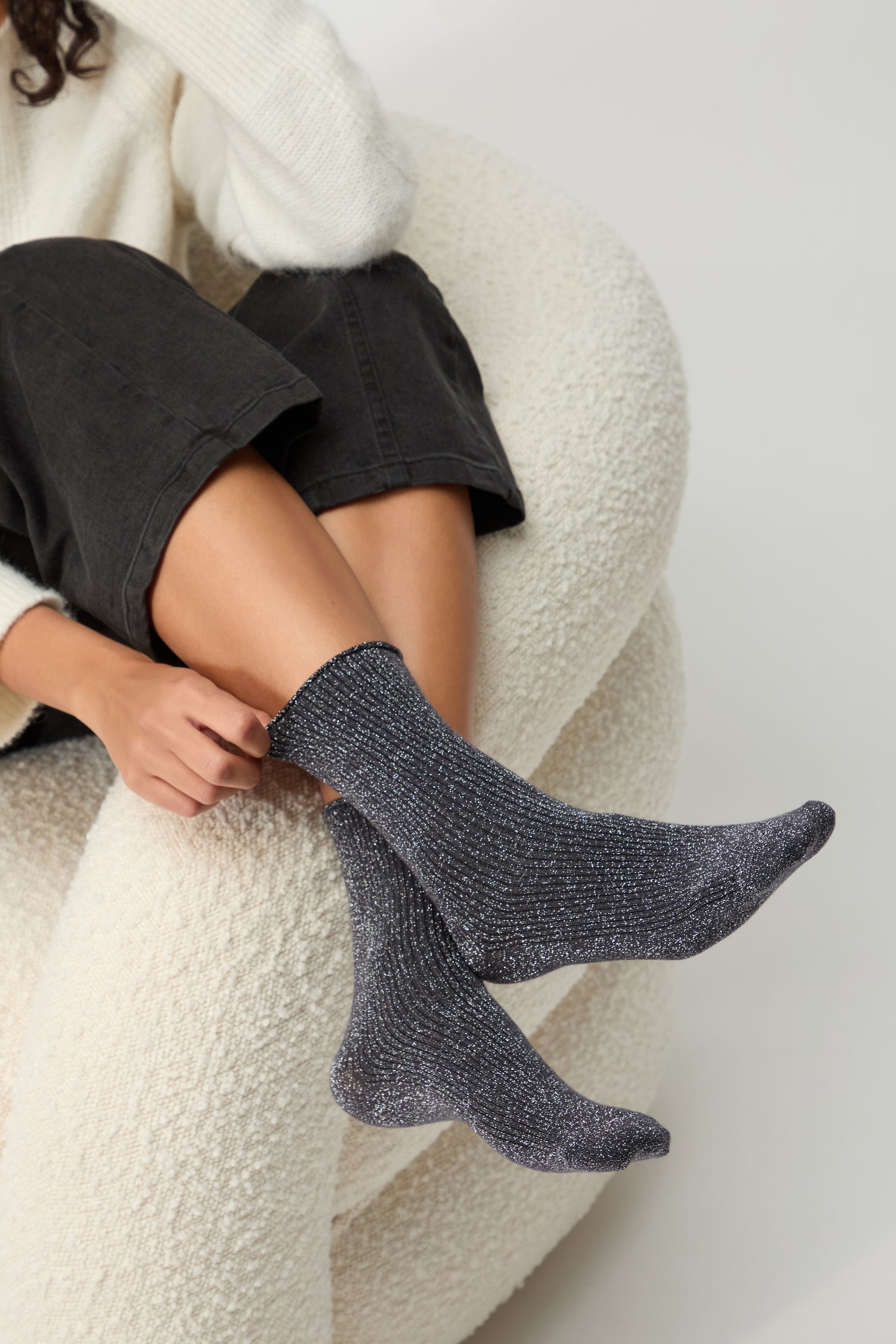 Person seated on a textured white chair, wearing Bamboo Sparkle Dark Grey Socks, black pants, and a cream sweater.