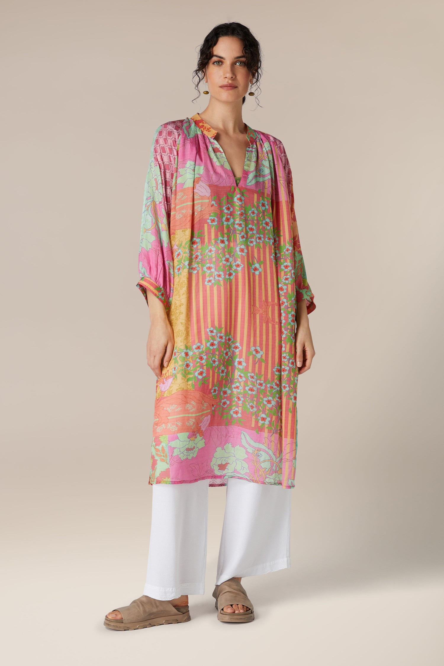 Woman wearing a colorful Geometric Floral Silk Beach Dress, paired with white trousers and sandals, standing against a neutral background.