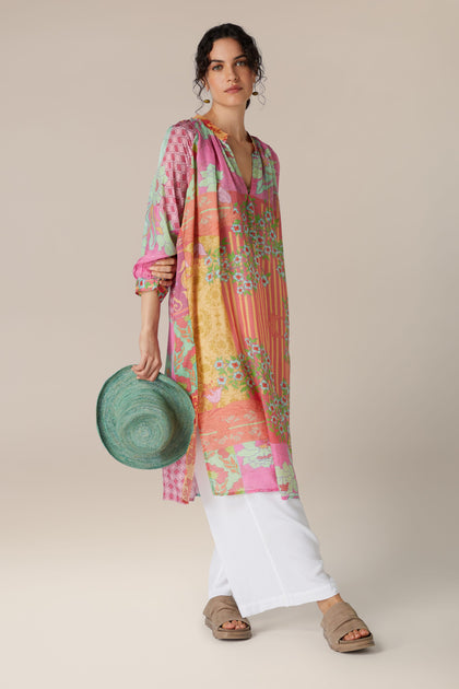 A woman in a Geometric Floral Silk Beach Dress and white trousers holding a green sun hat.