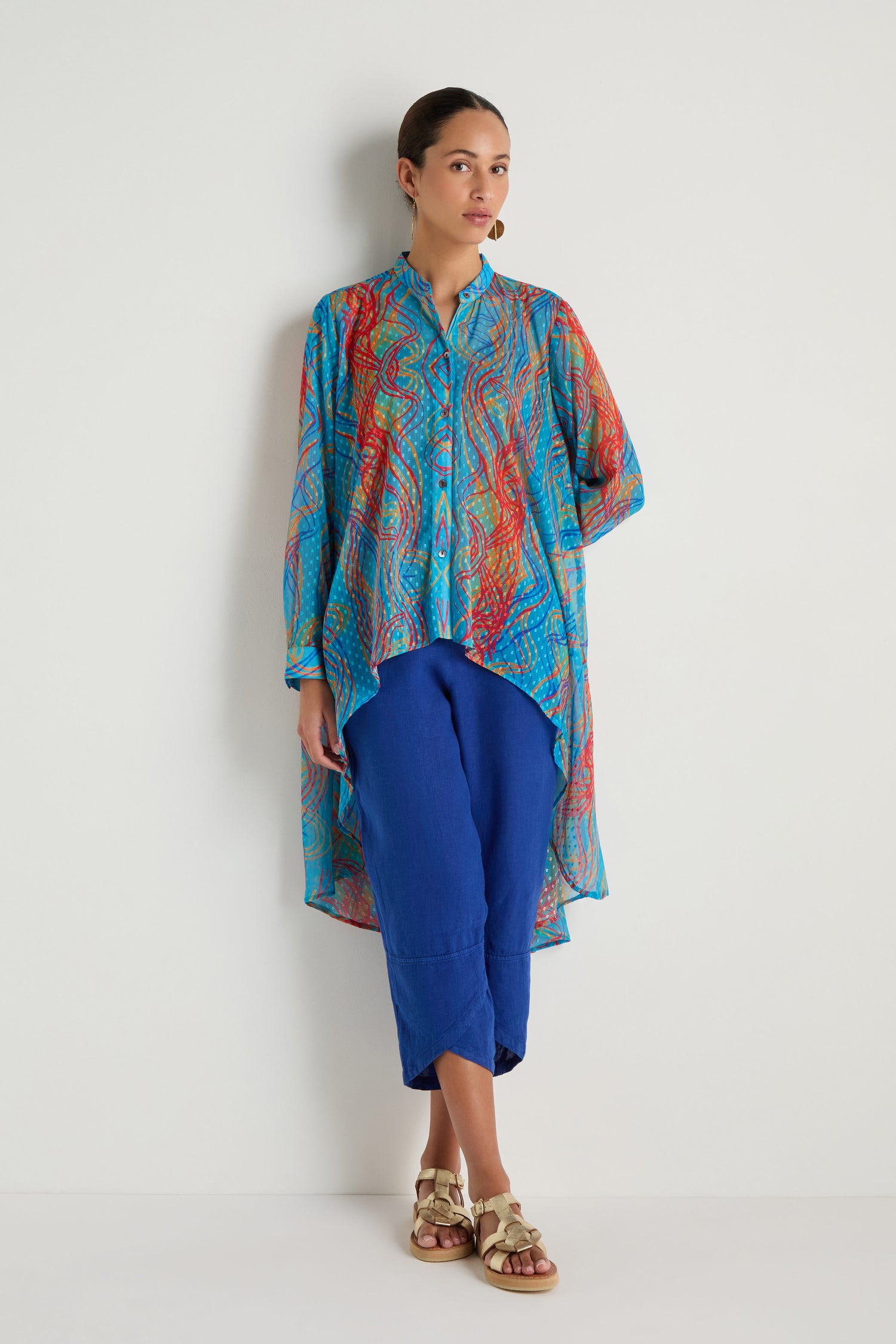 A woman stands against a white wall wearing the Twirl Print Dobby Shirt with a hi-lo hem over blue pants and gold sandals. Her hair is tied back and she looks forward.