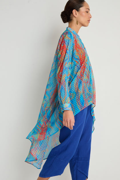 Woman in profile wears the Twirl Print Dobby Shirt—a colorful, flowing tunic with a hi-lo hem in blue and red—over blue pants, set against a plain light background.