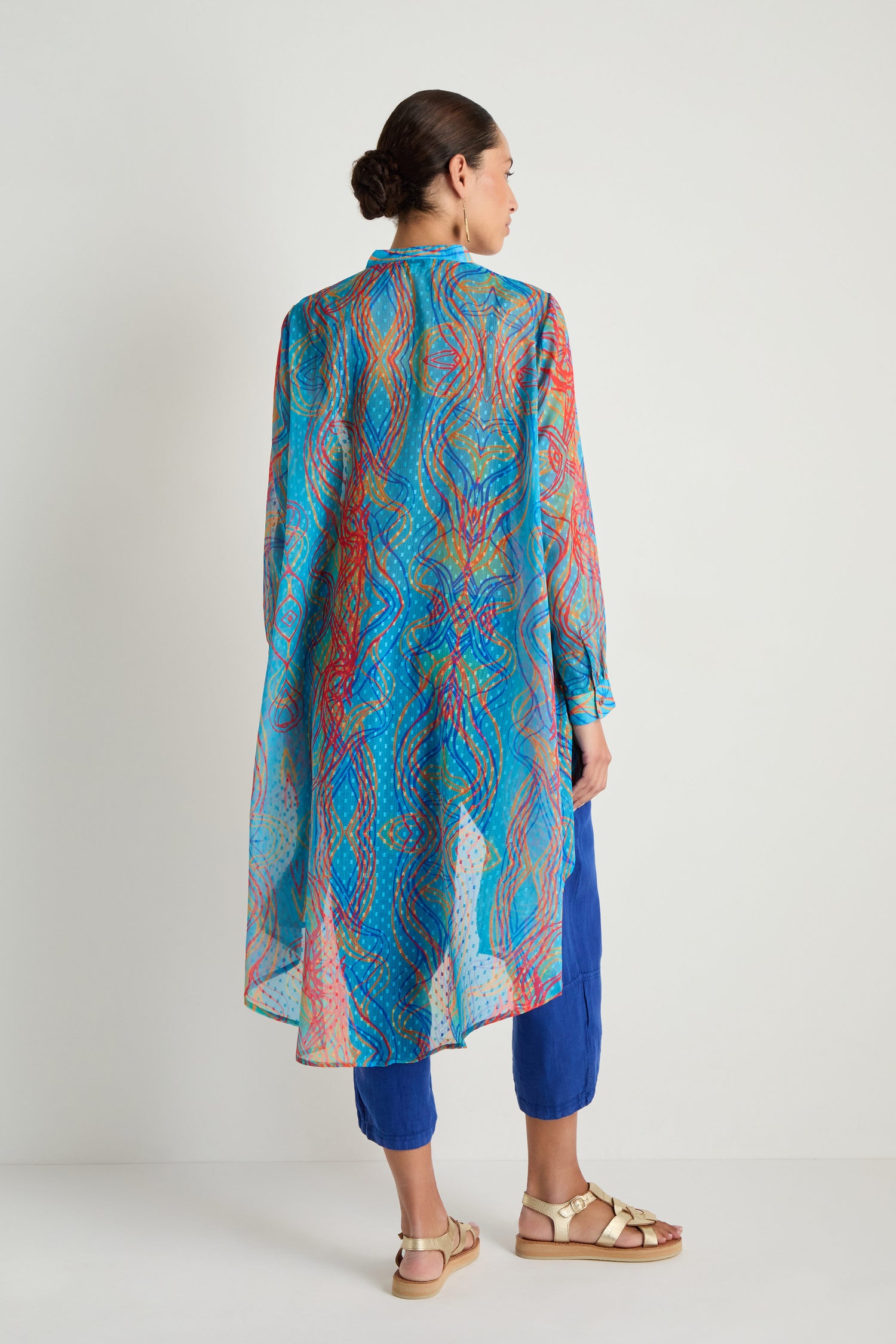 A woman stands with her back to the camera, wearing the Twirl Print Dobby Shirt—a colorful sheer tunic with a hi-lo hem—over blue pants and beige sandals, styled as a perfect lightweight layer against a plain background.