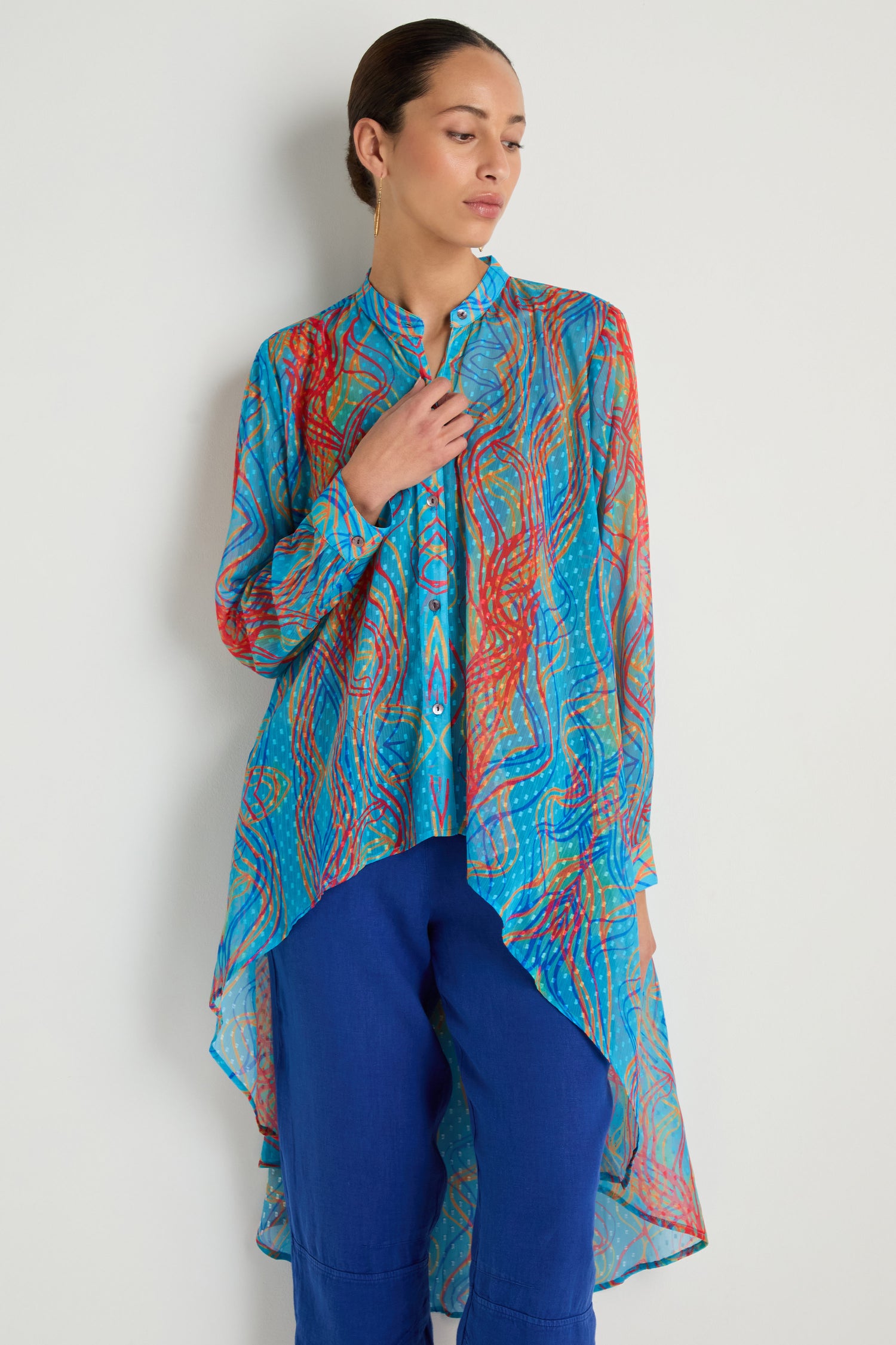 A woman stands against a plain wall wearing the Twirl Print Dobby Shirt, a long asymmetrical tunic with a hi-lo hem and multicolored patterns, styled over blue pants.