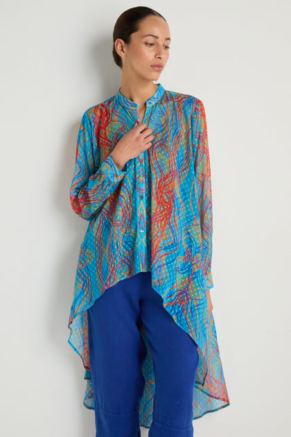A woman stands against a plain wall wearing the Twirl Print Dobby Shirt, a long asymmetrical tunic with a hi-lo hem and multicolored patterns, styled over blue pants.
