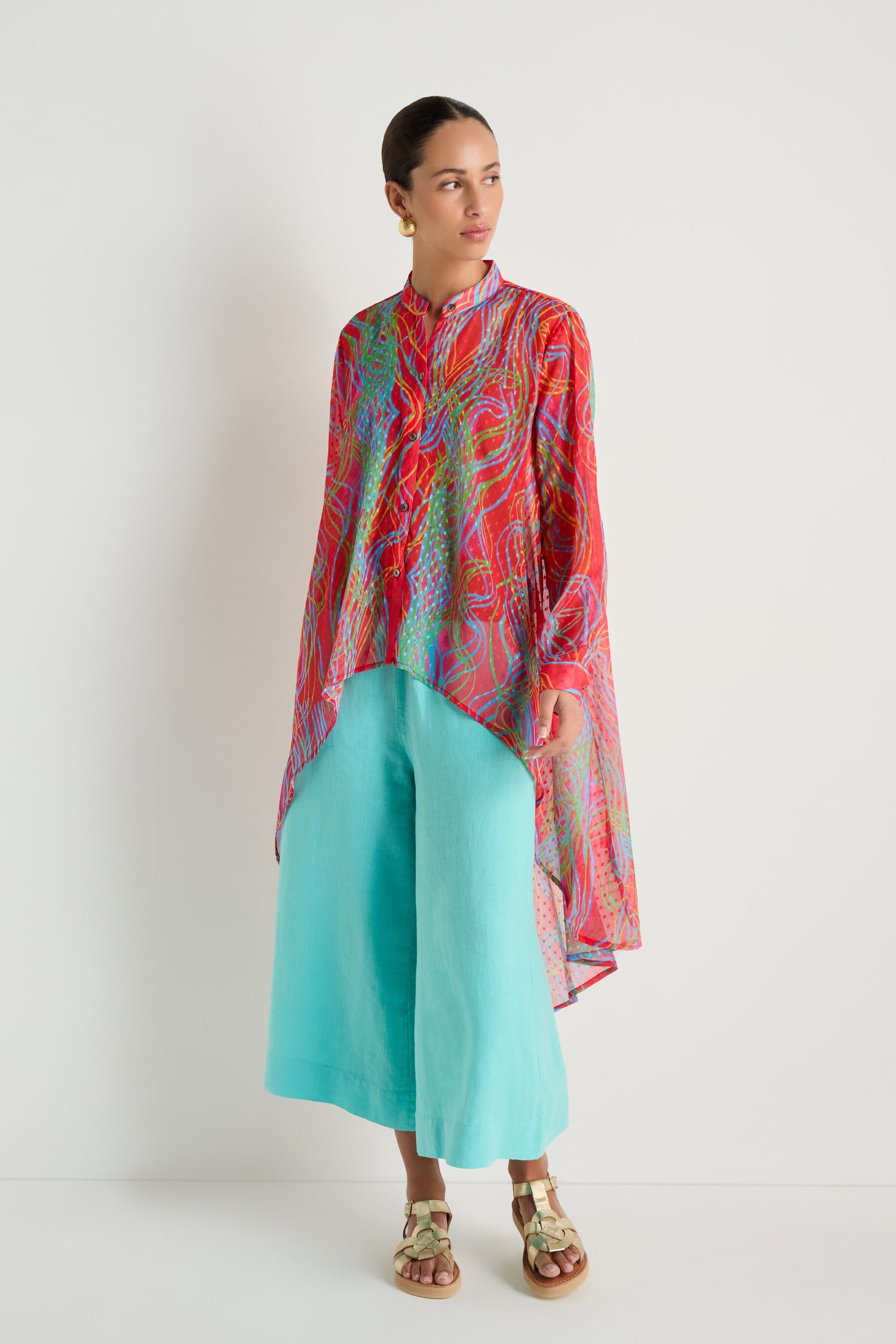 A person stands against a white background wearing the Twirl Print Dobby Shirt—red with patterns and a hi-lo hem—paired with turquoise wide-leg pants and gold sandals.