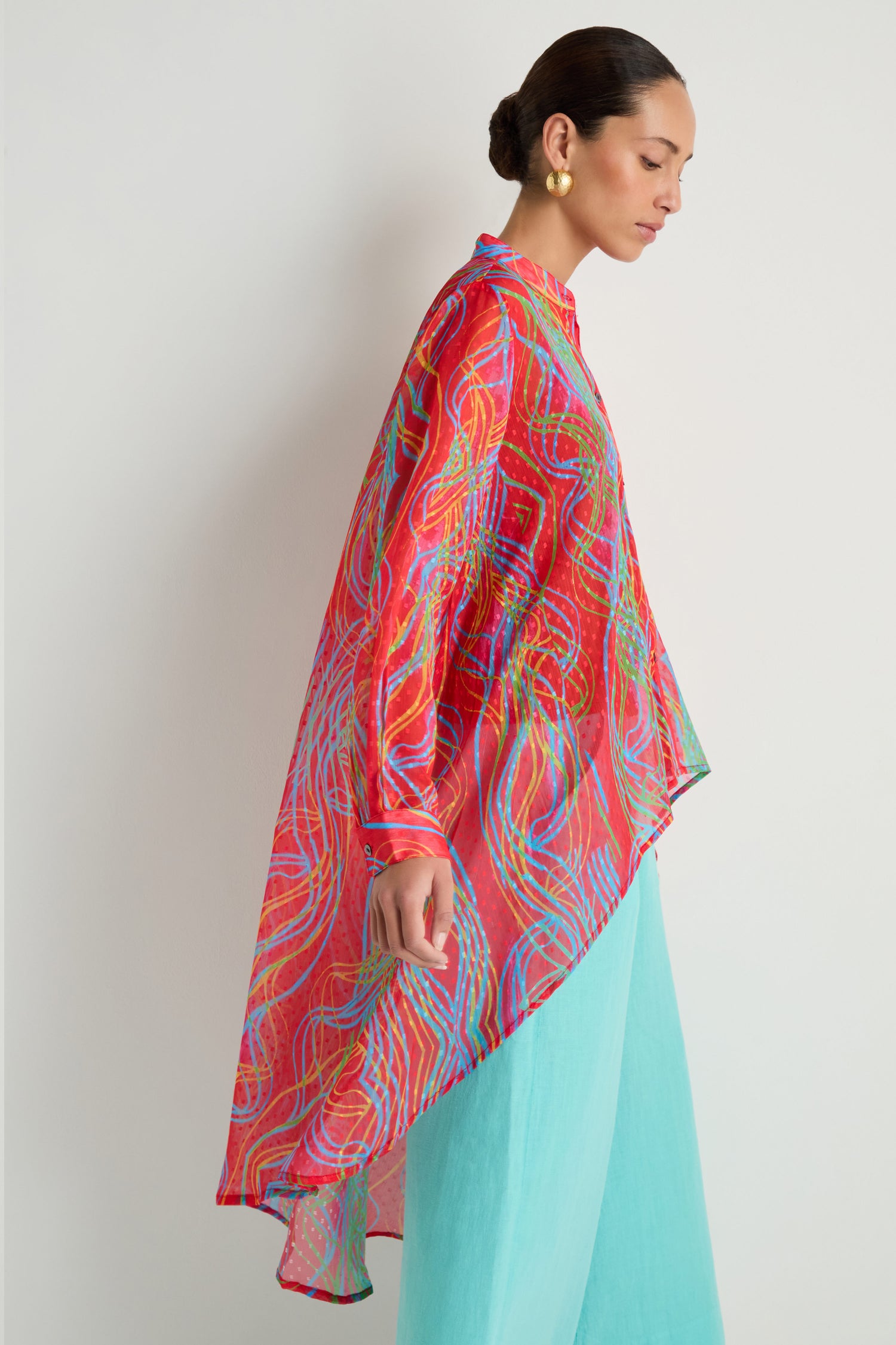 A woman in profile wears the Twirl Print Dobby Shirt, a sheer, brightly patterned asymmetrical tunic with a hi-lo hem over light blue pants, her hair in a low bun and gold earrings.