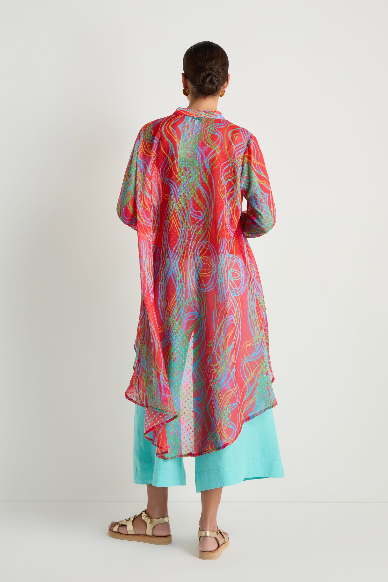 Person stands with back to camera, wearing the Twirl Print Dobby Shirt featuring a long, colorful hi-lo hem, paired with light blue pants and beige sandals against a plain white background.