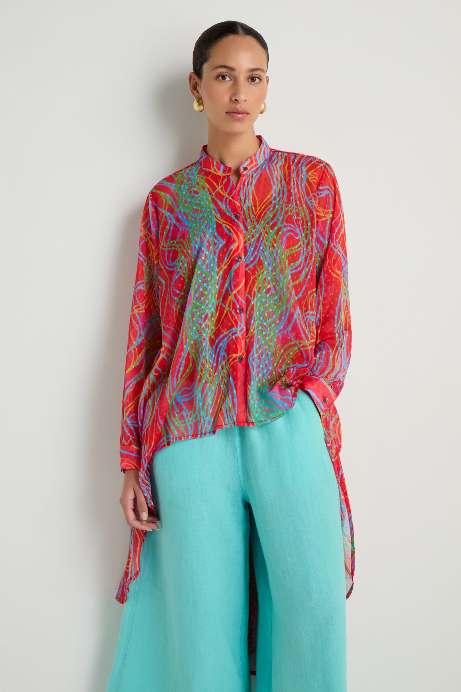 A woman wears the Twirl Print Dobby Shirt, a long-sleeved, multicolored red top with a hi-lo hem, paired with loose turquoise pants, standing against a plain light background.