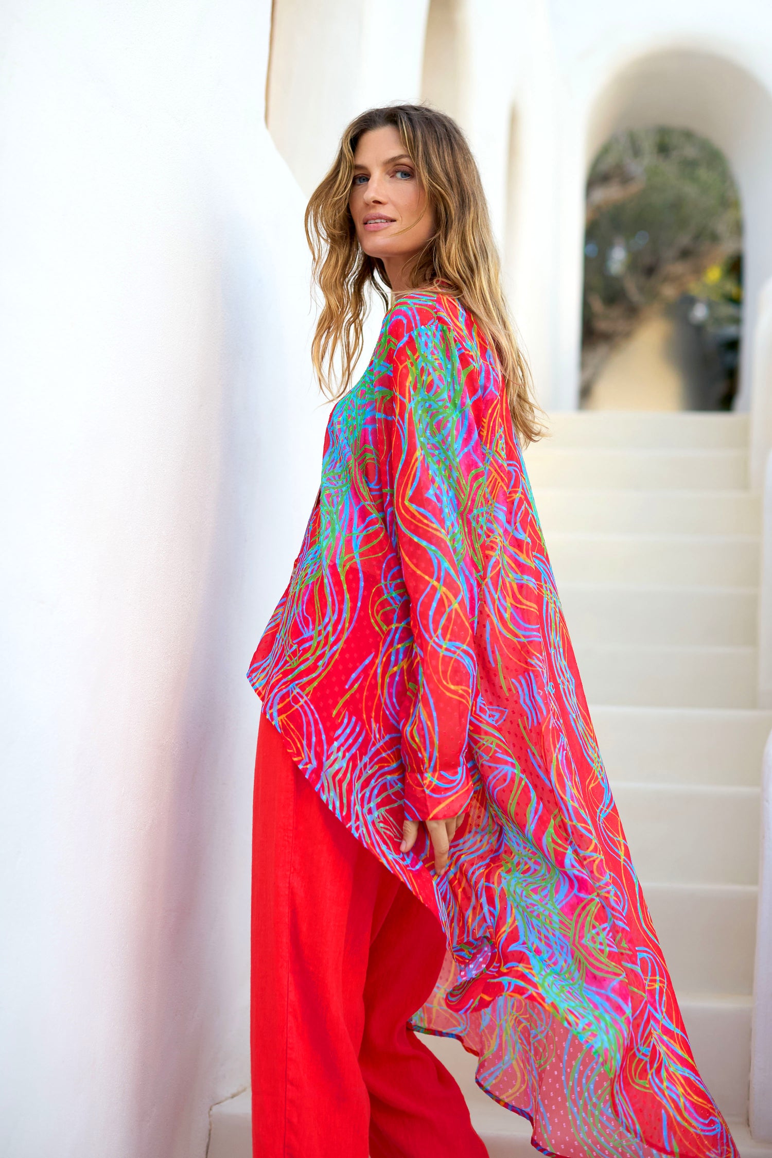 A woman stands on white stairs, wearing the Twirl Print Dobby Shirt—a bright red long top with blue and green patterns, semi-sheer fabric, and a hi-lo hem—as she looks over her shoulder towards the camera.