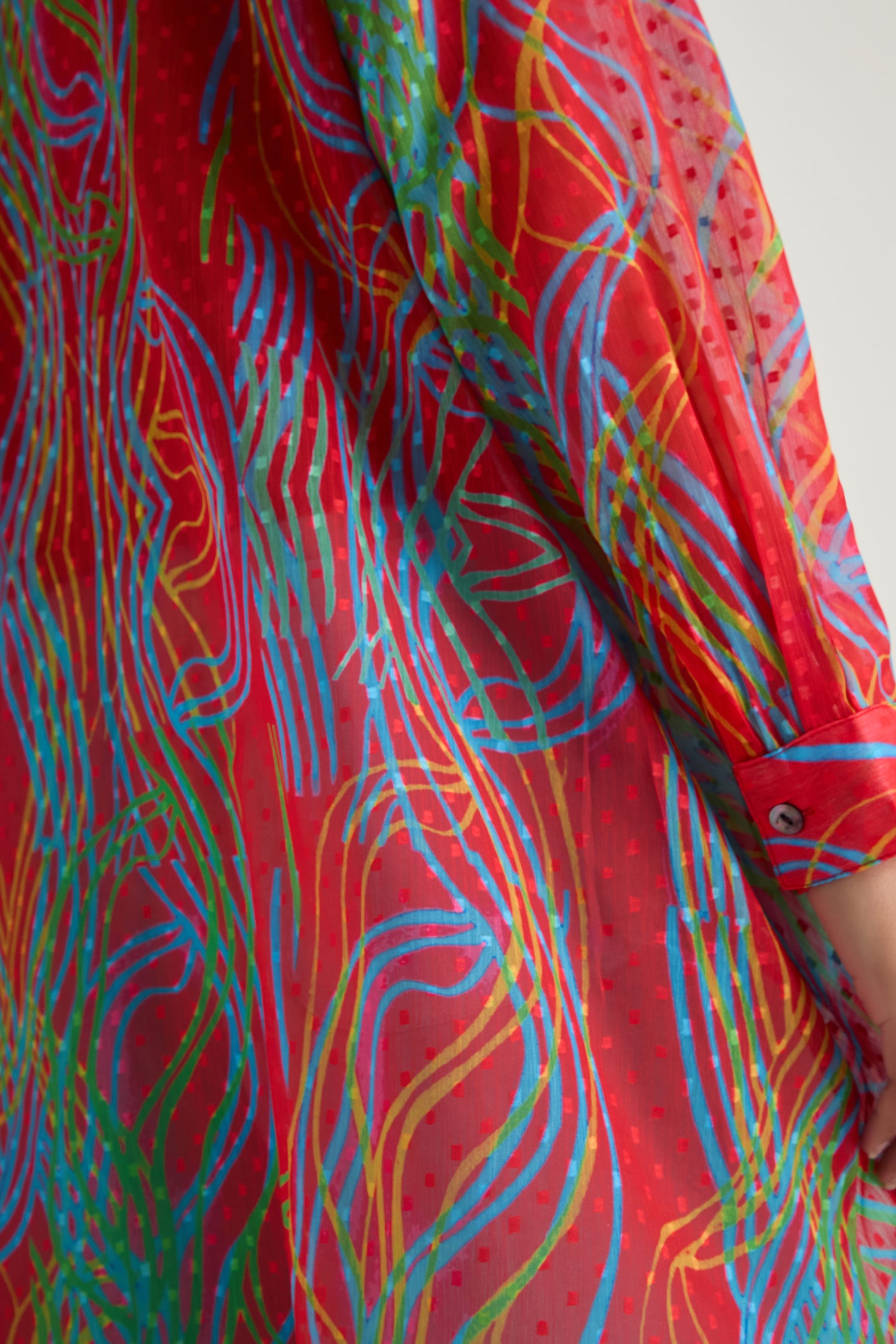 Close-up of an arm in the Twirl Print Dobby Shirt, a red lightweight layer with abstract wavy blue, green, and yellow lines plus dotted patterns. Features a cuff with button for added style.