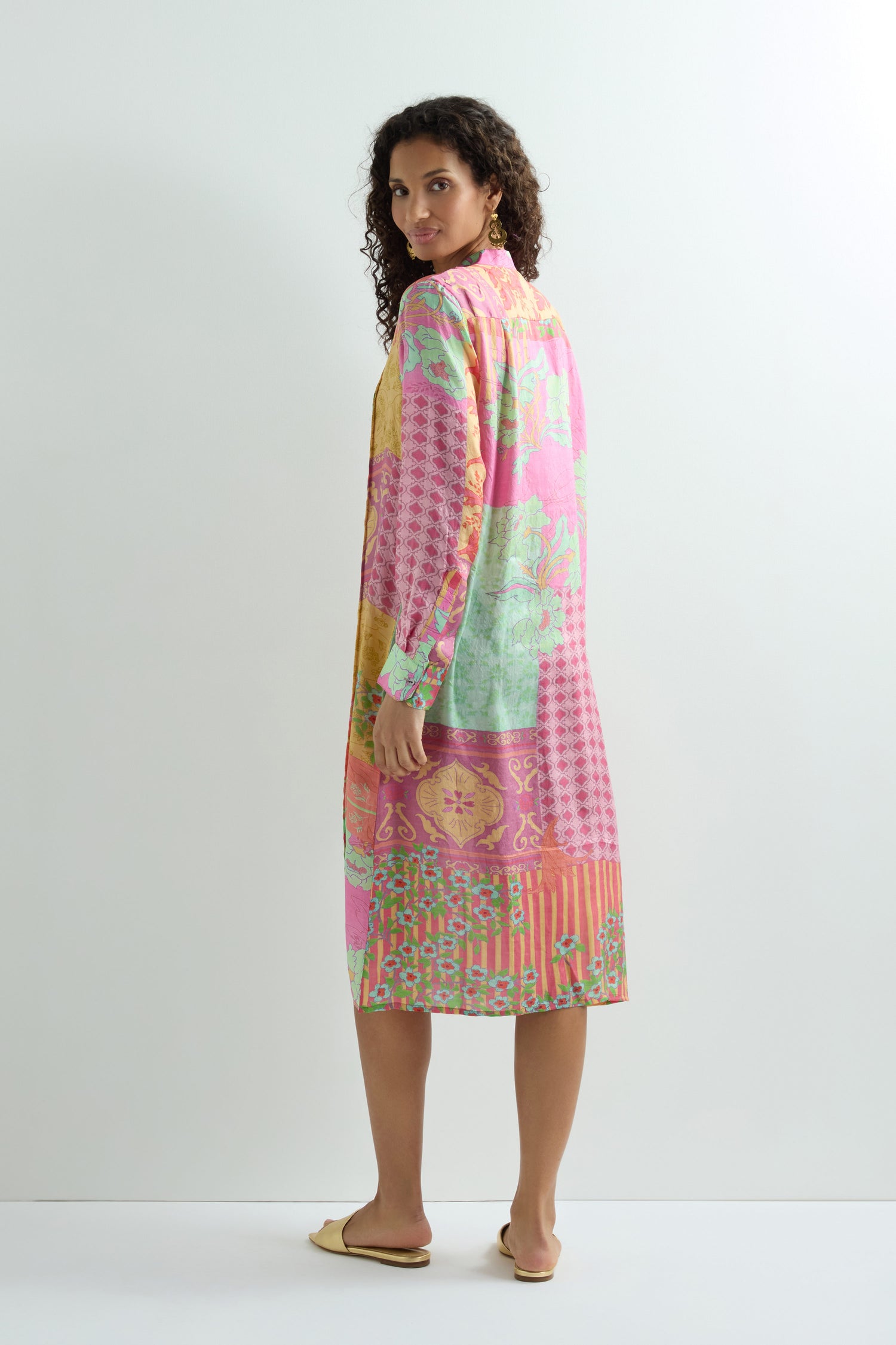 A person stands with one foot forward, adorned in a vibrant Geometric Floral Silk Shirt Dress featuring a lively mix of pink, green, and yellow patterns.