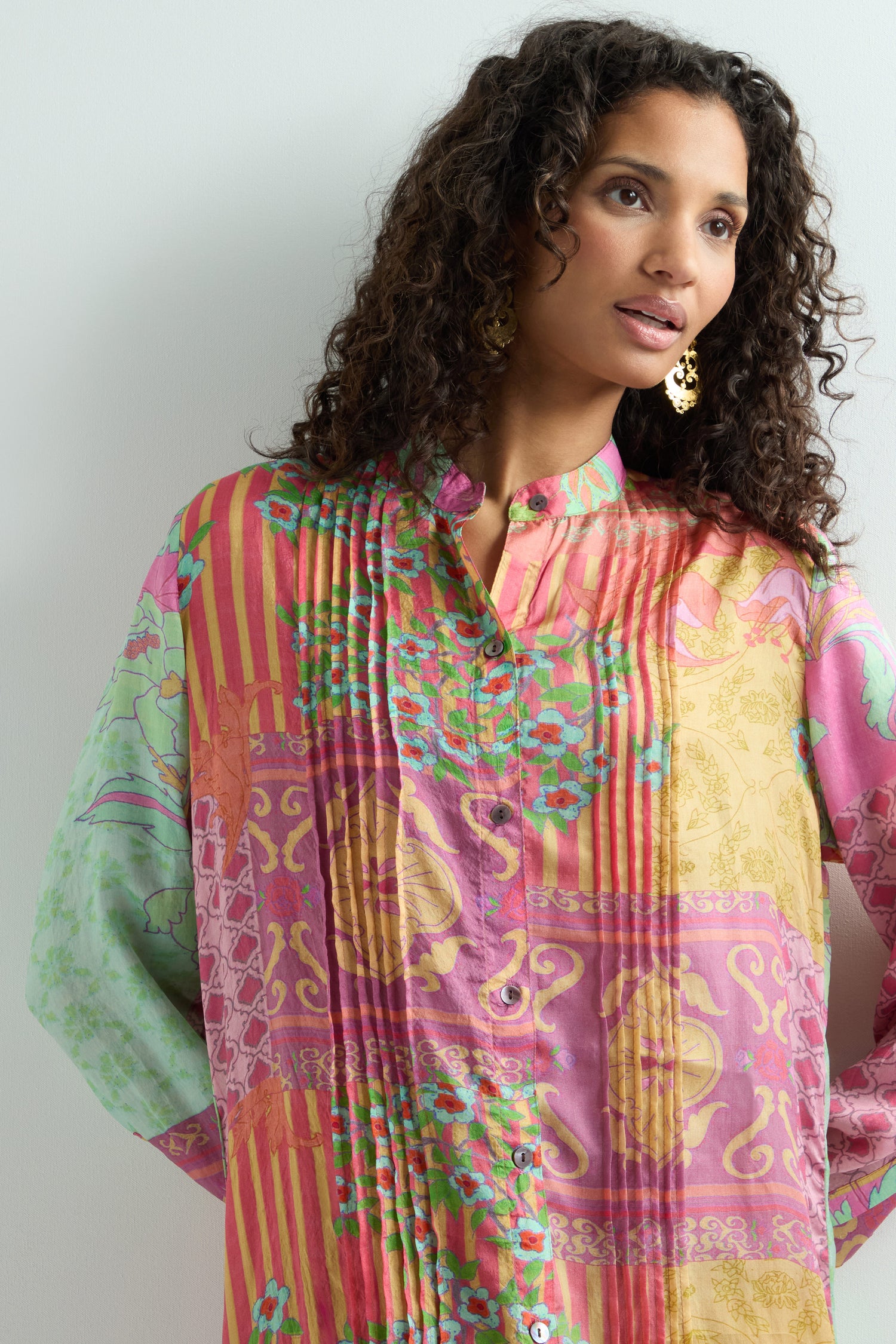 A woman in a vibrant Geometric Floral Silk Shirt Dress with long sleeves and intricate floral designs stands against a neutral background. She has long, curly hair and wears gold hoop earrings.
