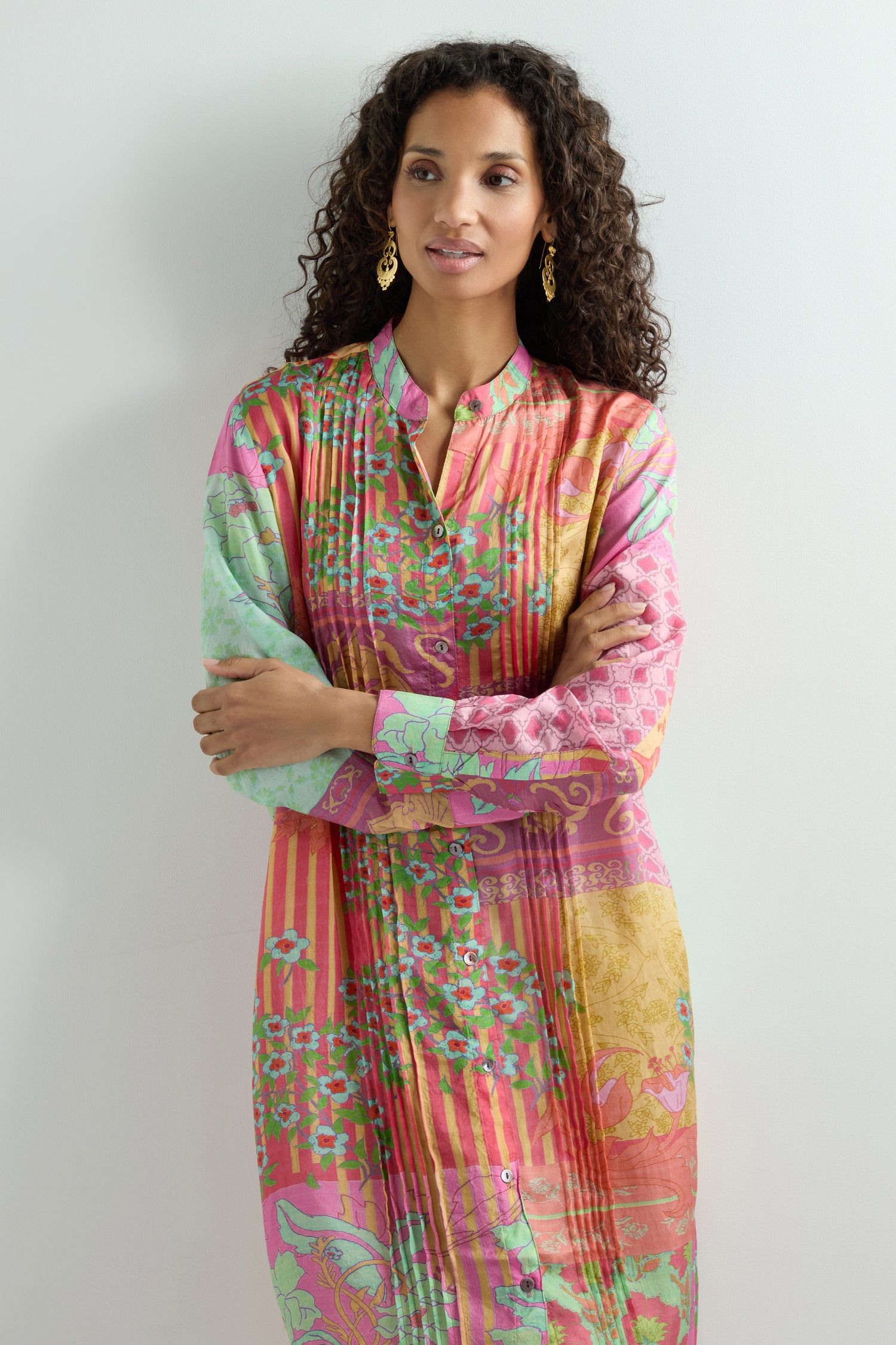 A woman with curly hair stands confidently against a plain background, her arms crossed. She wears a vibrant Geometric Floral Silk Shirt Dress paired with gold earrings that catch the light.