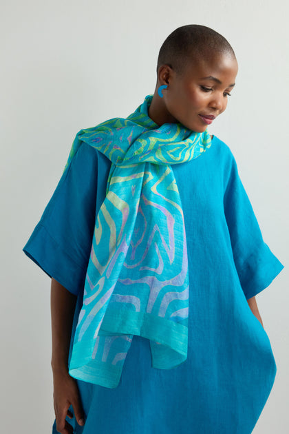 A person in a blue dress, wearing the luxurious Ripple Silk Scarf with an abstract teal pattern, stands against a plain background, gazing slightly downward.