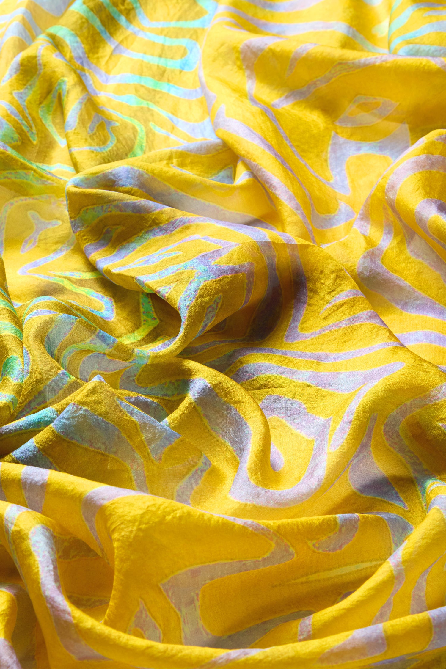 The Ripple Silk Scarf, with its yellow silk fabric and swirling turquoise and white patterns, radiates vibrant energy while offering a textured and layered look.