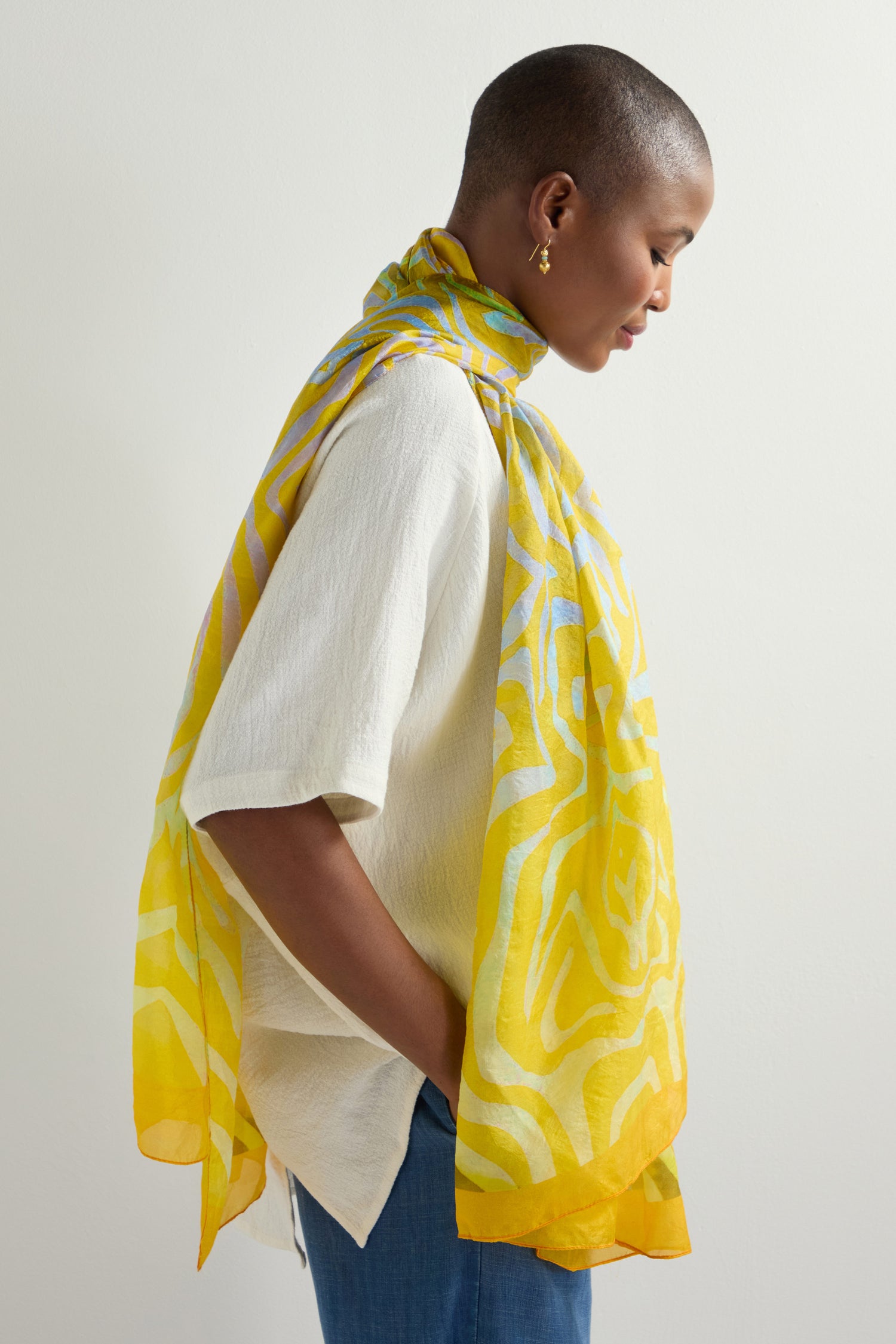 In profile, a person radiates vibrant energy with short hair and hoop earrings, adorned in a yellow Ripple Silk Scarf, textured white shirt, and jeans against a plain background.