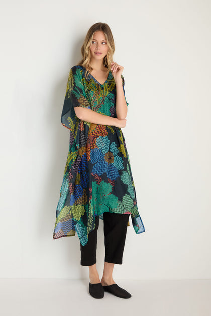 A woman stands against a plain background wearing the Pixelated Petals Silk V-Neck Kaftan, featuring a vibrant pixelated floral print, paired with black pants and black slip-on shoes.