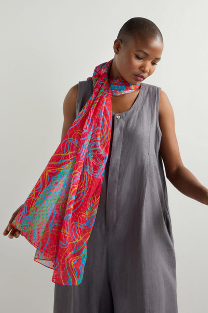 Wearing a sleeveless gray dress, a person stands against a plain background with a Twirl Print Dobby Scarf gracefully draped over their shoulders, its distinct pattern adding flair.