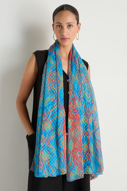 A woman in a sleeveless black dress stands against a plain wall, wearing the Twirl Print Dobby Scarf with blue and red abstract swirls, paired with gold hoop earrings.