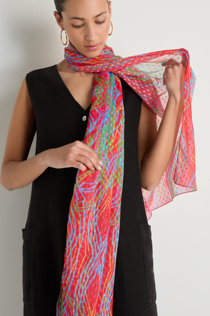 A person in a sleeveless black dress adjusts the Twirl Print Dobby Scarf, featuring red, green, and blue wavy lines with small dots in an abstract swirl design.