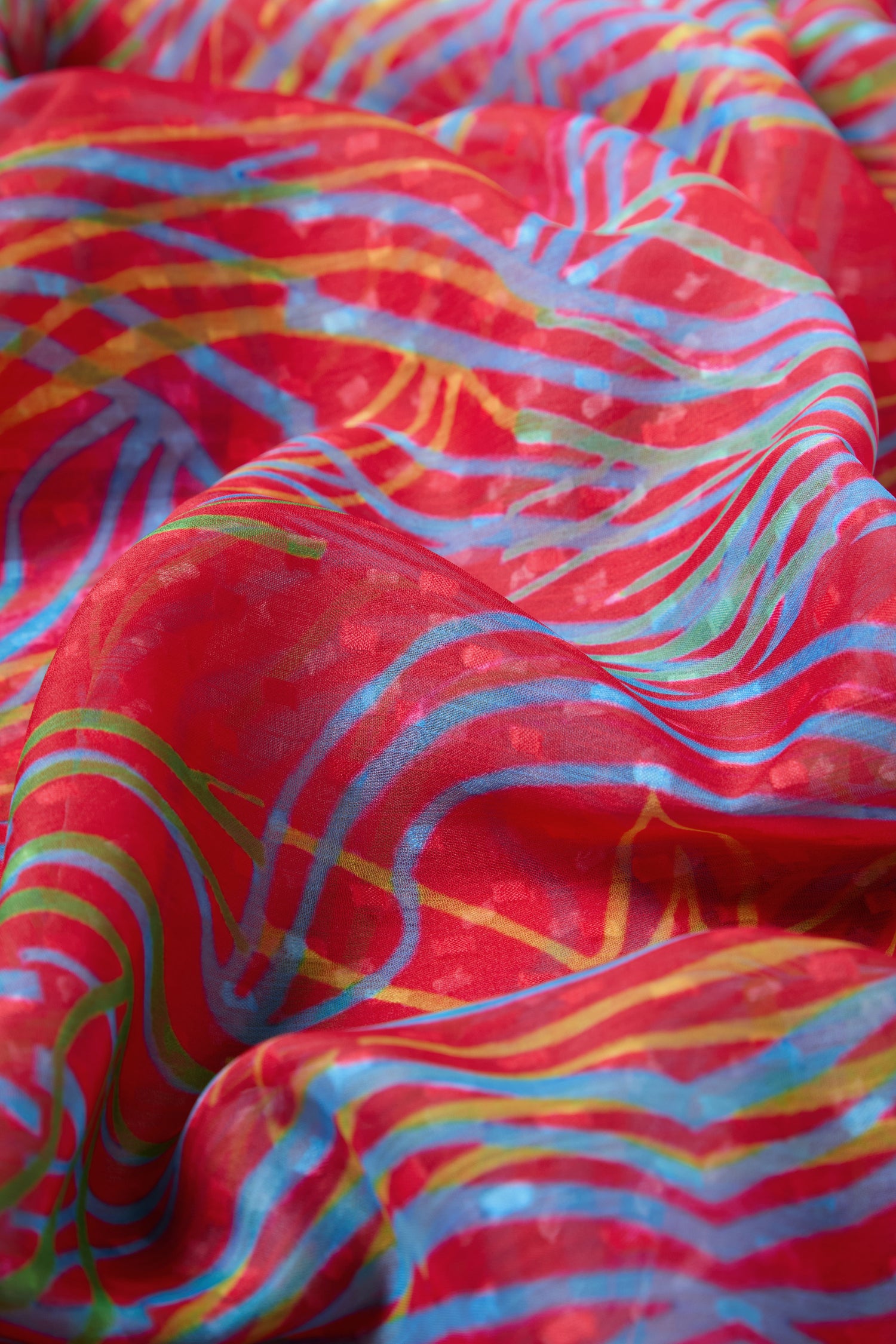 The Twirl Print Dobby Scarf features red fabric with blue, yellow, and green abstract wavy lines in a swirl pattern, elegantly draped to highlight its textured folds.